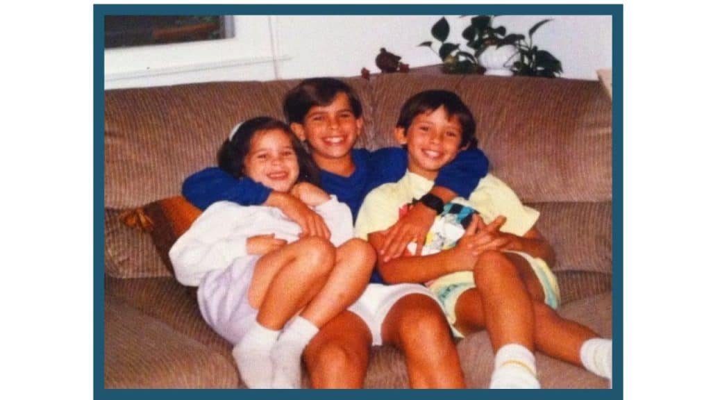 Jessica, Nick, and Alex Ortner as children sitting on a brown couch with arms around each other.